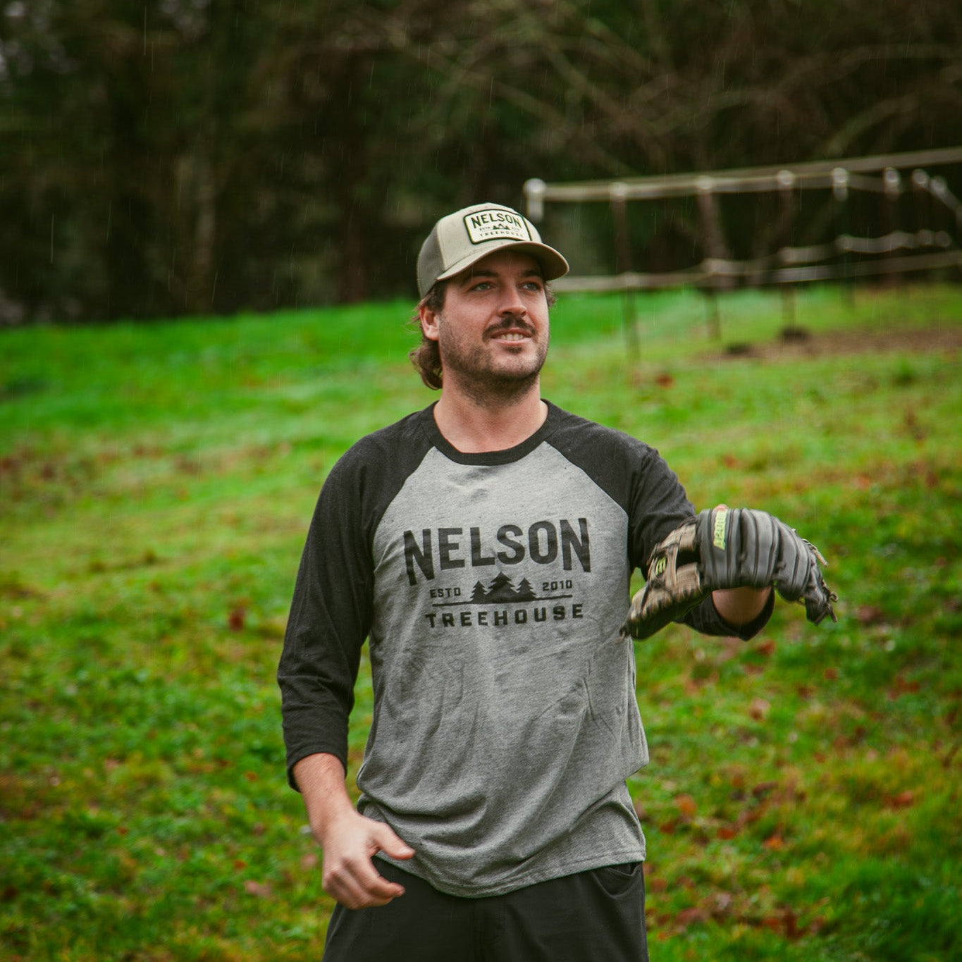 Man wearing a gray and black baseball shirt with 'Nelson Treehouse' printed on it, standing in a grassy field.