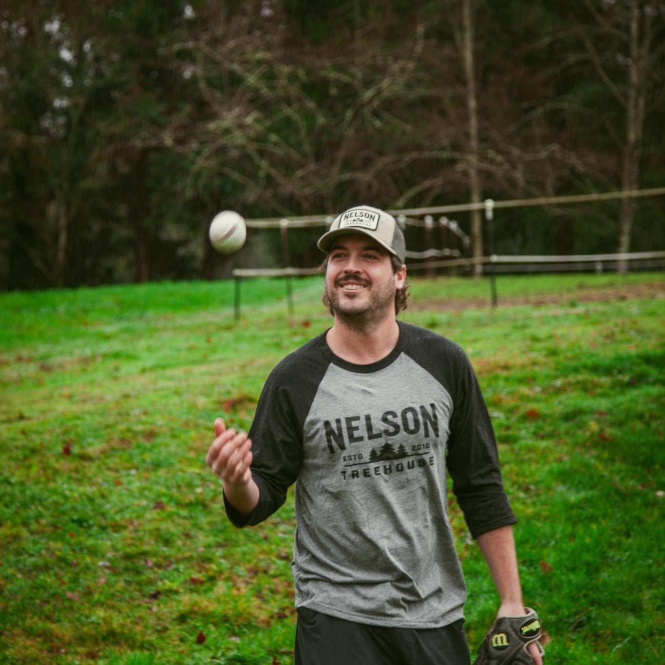 Man in a gray 'Nelson Treehouse' shirt holding a baseball and glove on a grassy field.